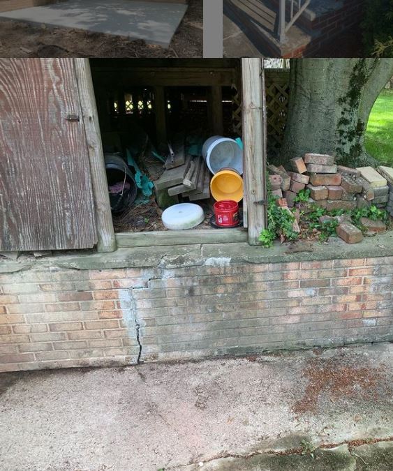 Cracked brick foundation with an open shed door revealing buckets and debris inside. A tree trunk is visible on the right.