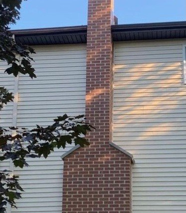 Brick chimney against a beige-sided house, with sunlight casting shadows. A tree's leaves partially frame the left side.