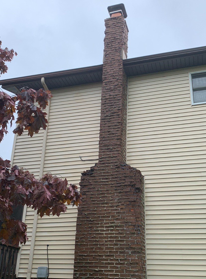 Brick chimney on the side of a two-story house; it is weathered and in need of repair. The sky is overcast.