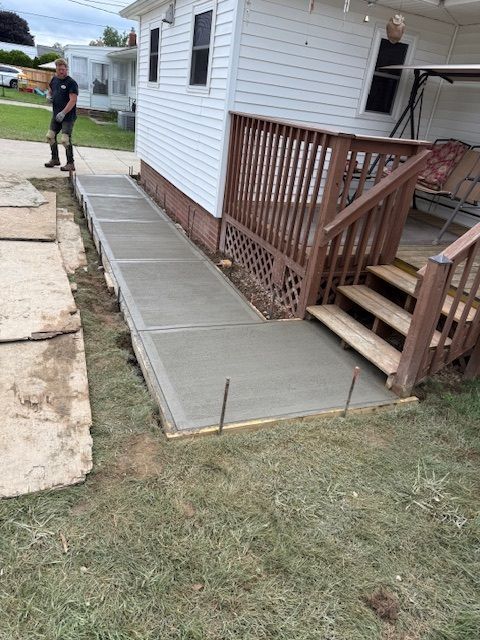 Newly poured concrete walkway running along a house and wooden deck, with a person standing on a lawn nearby.