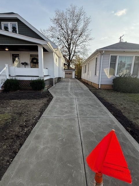 A concrete driveway runs between two white houses, leading toward a distant garage under a bare tree, with a red cone.