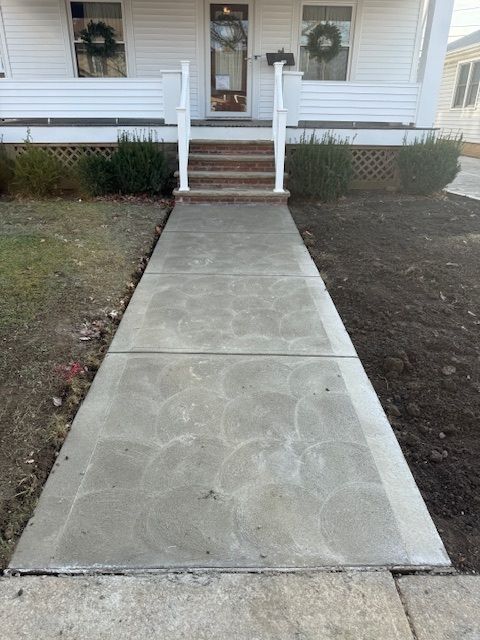 A concrete walkway leads up to the front porch steps of a white house with wreaths on the windows.