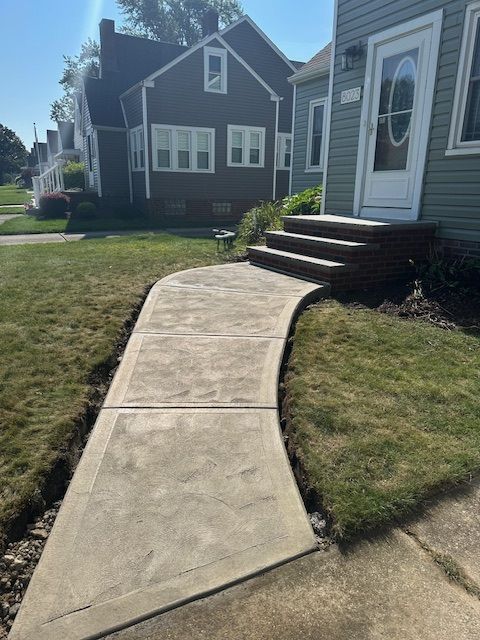 A new stamped concrete walkway leads to the front porch steps of a grey house on a sunny day.