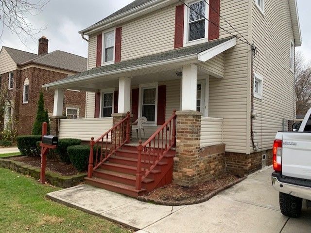 Two-story beige house with red shutters, steps, and trim. Brown brick accents at the base of the porch.