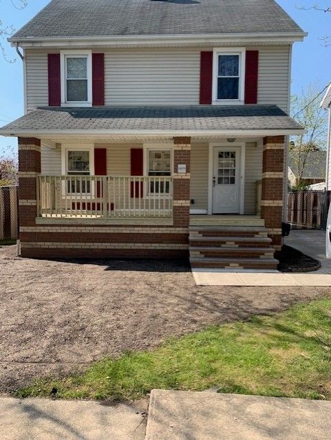 Two-story house with tan siding, red shutters, and a porch with brick columns. A front yard of dirt and grass.
