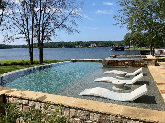 Infinity custom pool and spa with in-water lounge chairs overlooking a scenic lakefront view, surrou