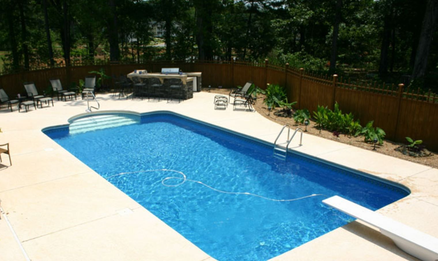 Rectangular backyard pool with diving board, outdoor kitchen, and lounge chairs surrounded by trees.