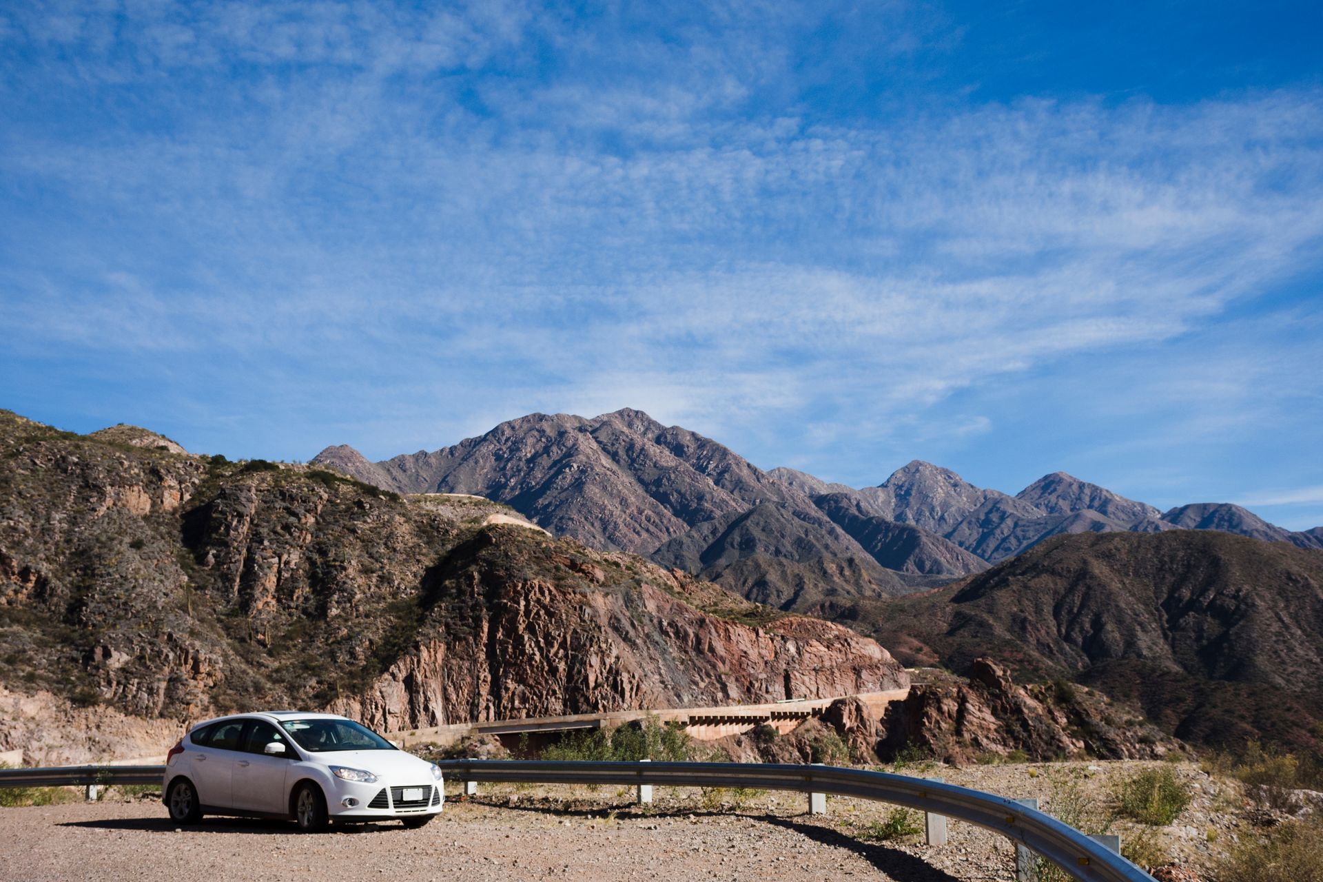 A white car is parked on the side of a road with mountains in the background.