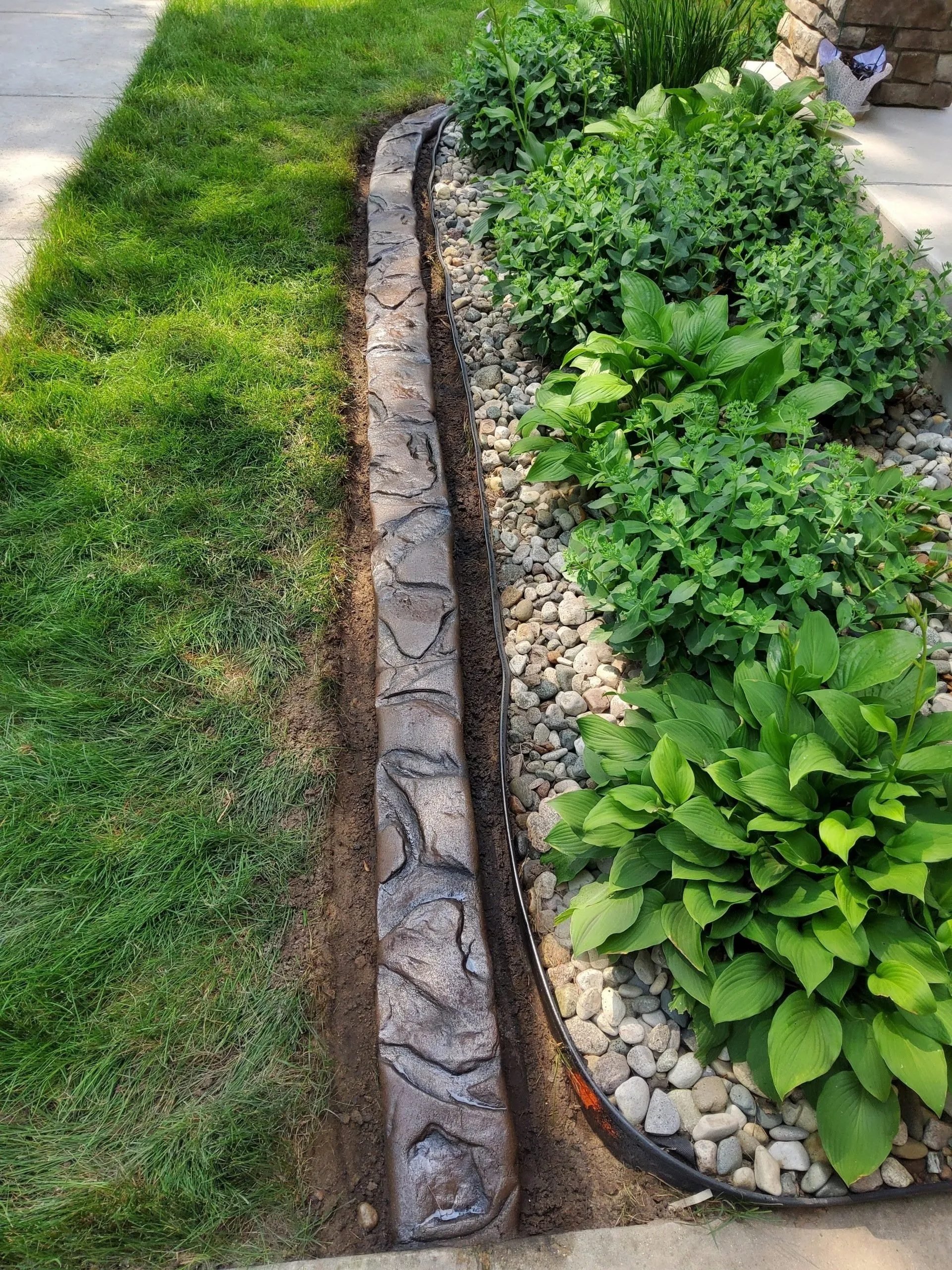 A decorative stone-look concrete landscape border separates a grassy lawn from a rock garden with leafy green plants.