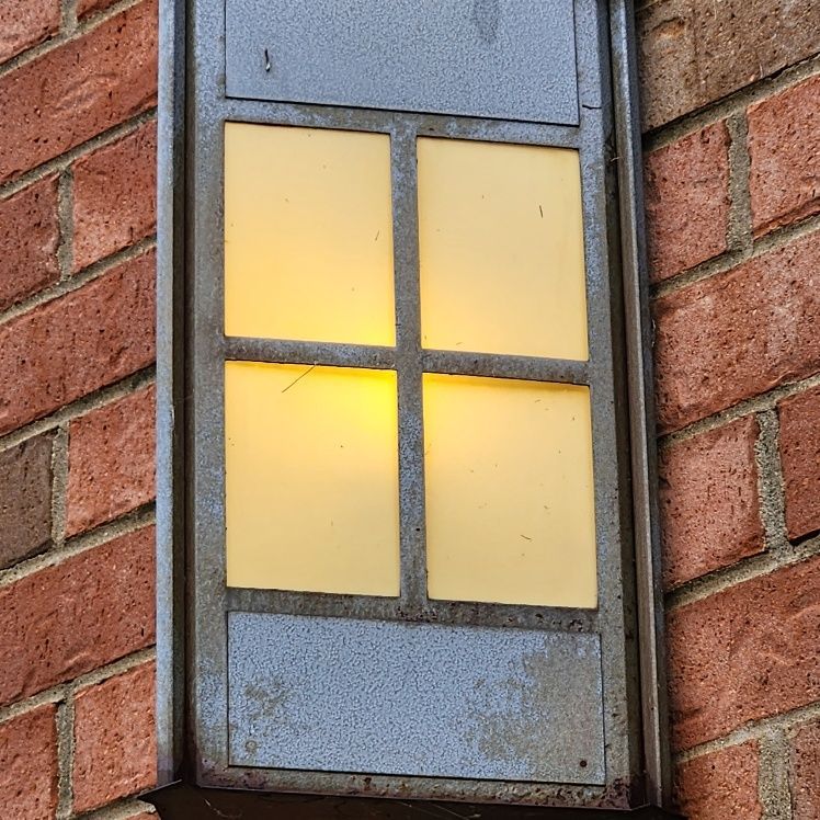 Gray rectangular outdoor light fixture on red brick wall; lit with yellow light.