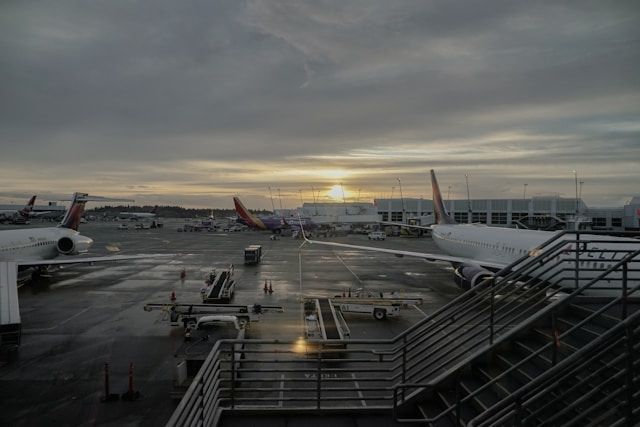 Airport scene at dusk with planes, terminal, and stairs; cloudy sky with sun peeking through.