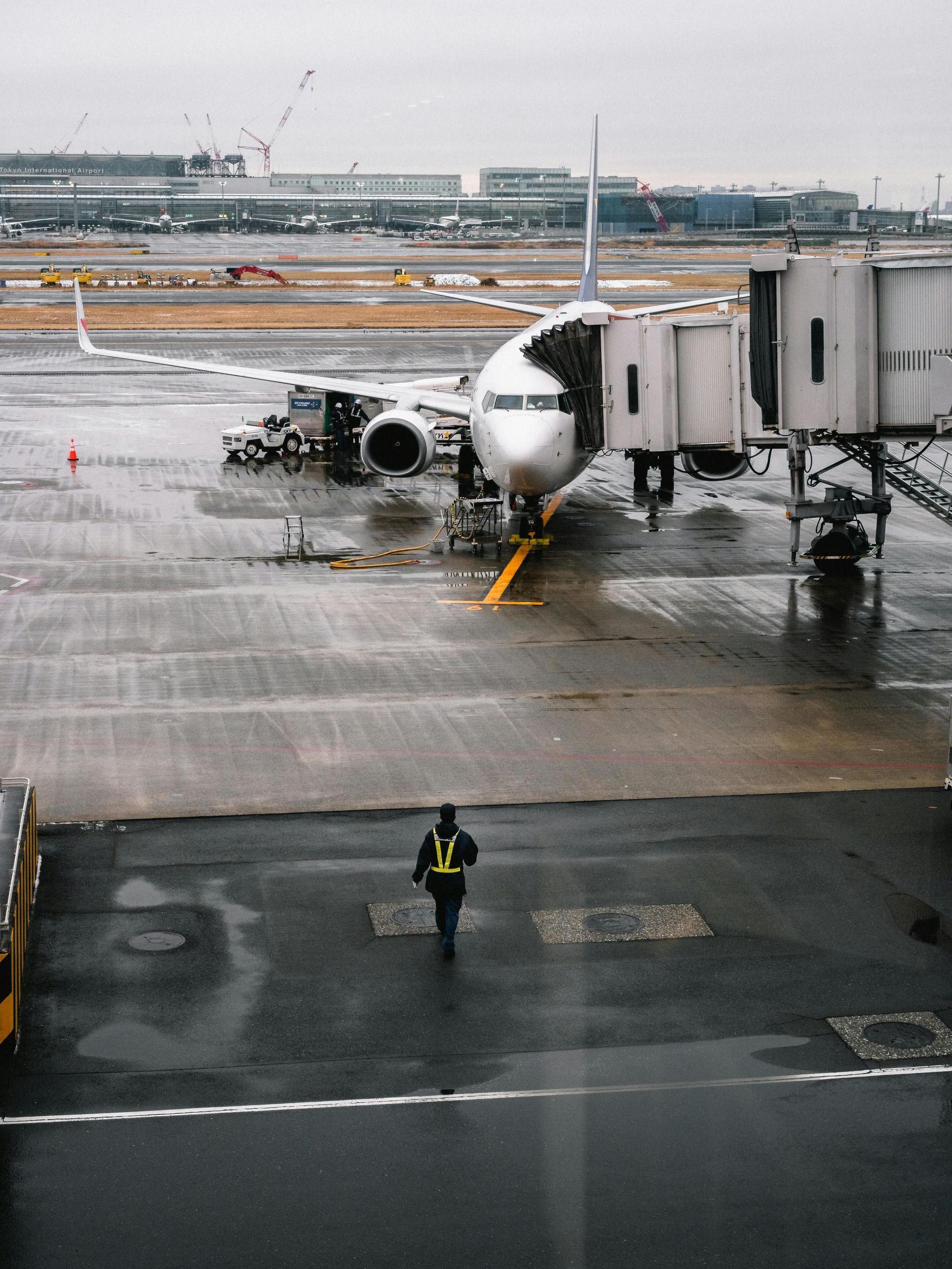 Airplane at a gate on a wet tarmac. A worker walks toward it. Gray sky, buildings in the background.