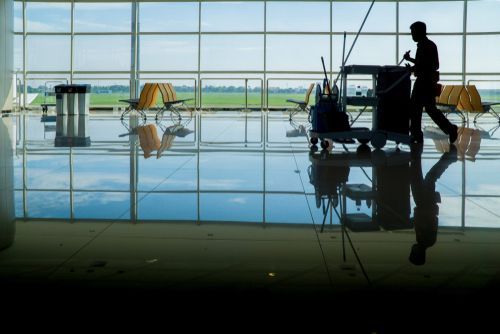 Silhouette of a person cleaning a reflective airport floor, trolley nearby, with outside view of fields.