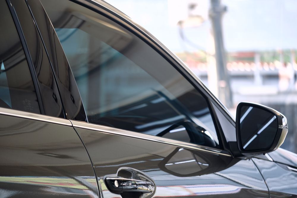 Close-up of a car's side, showing tinted window, side mirror reflecting bright lights, and door handle.