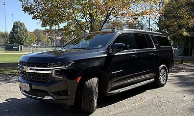 Black Chevrolet Suburban parked on pavement, with a park in the background.