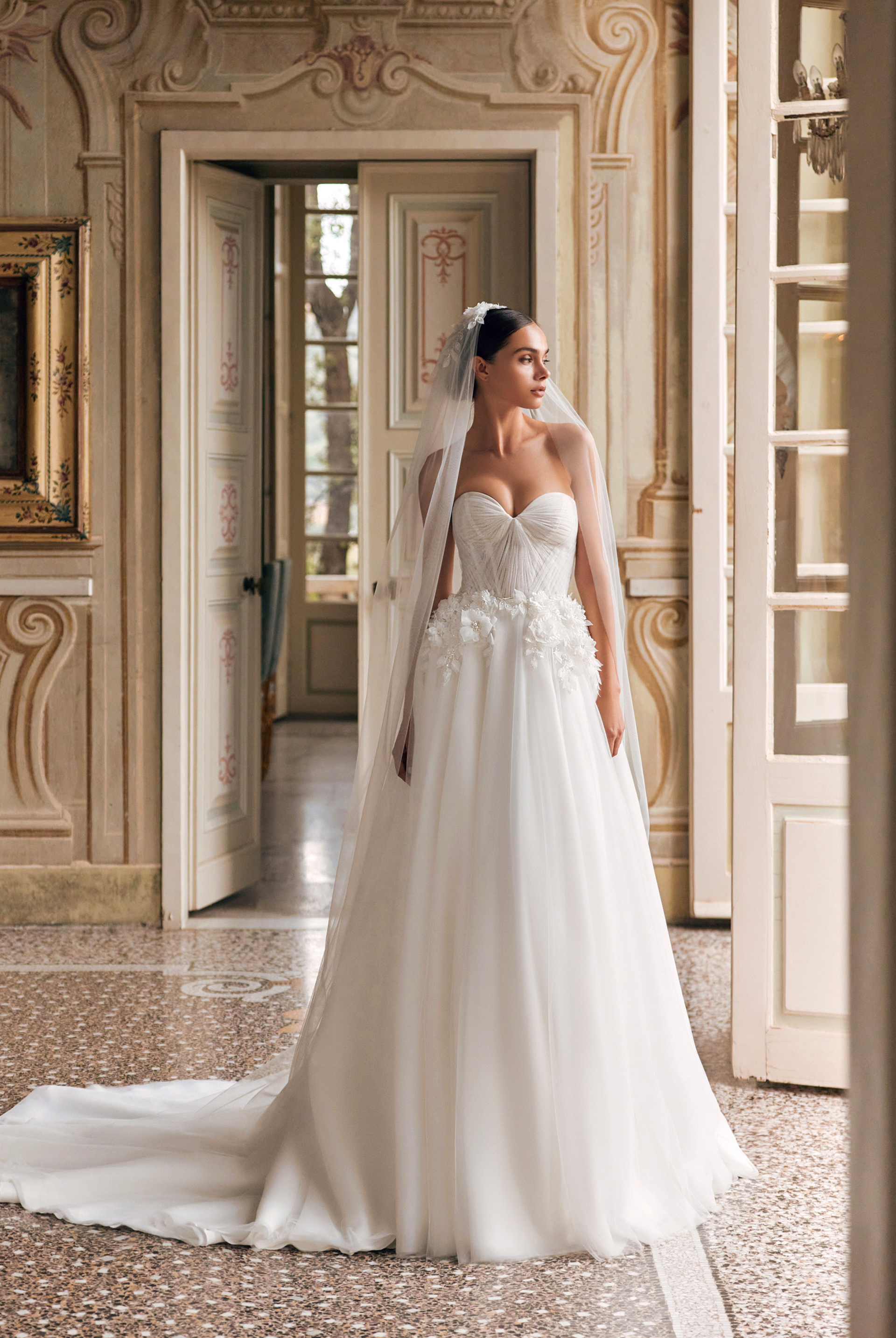 Bride in a white lace wedding gown standing in an ornate hallway with mirrored doors