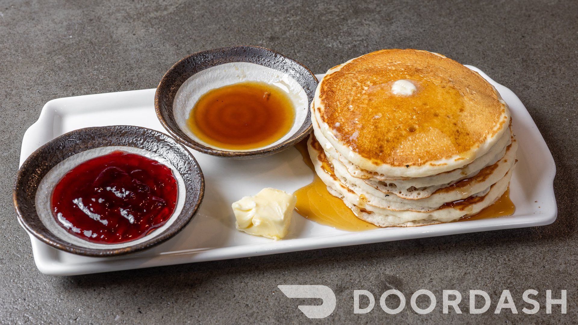 A stack of pancakes with syrup and jam on a white plate.