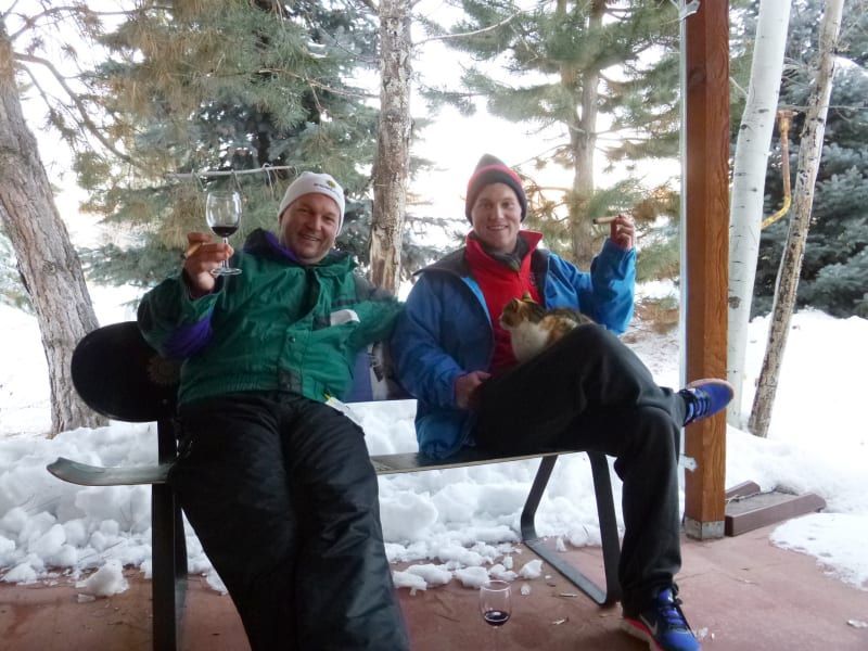 Two men are sitting on a bench in the snow toasting with wine glasses
