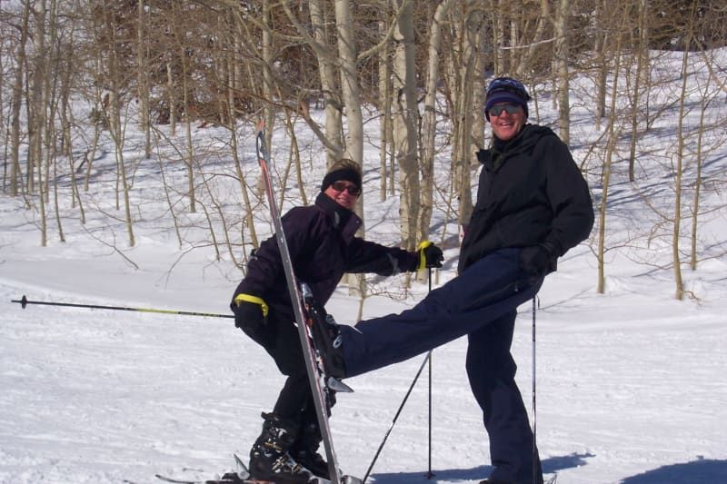 Two people are skiing in the snow and one of them is stretching his leg