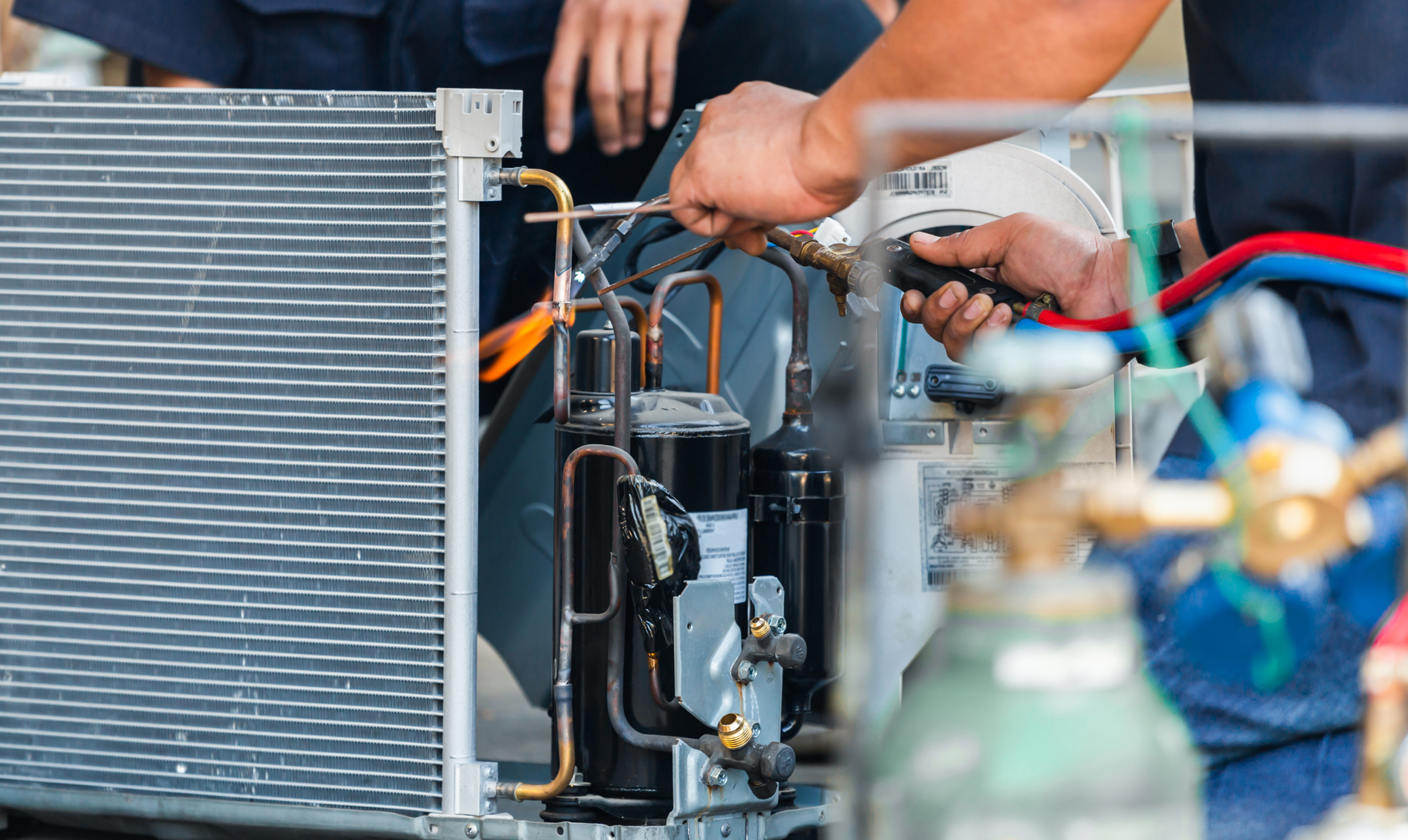 Person repairing an air conditioning unit; hands using tools, hoses, and copper tubing visible.