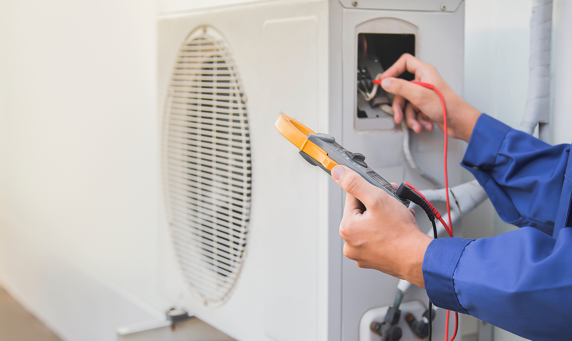 Technician in blue coveralls using a multimeter on an outdoor air conditioning unit.