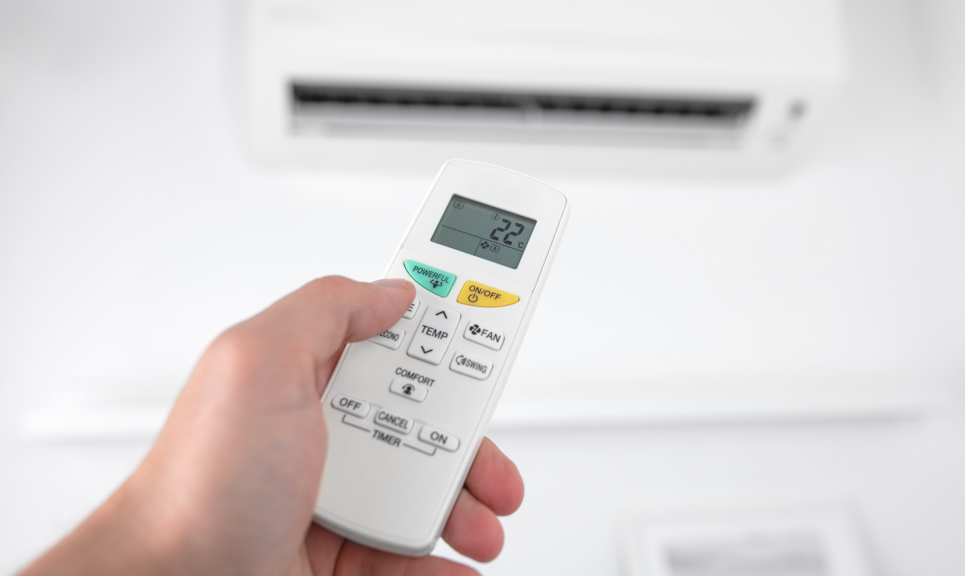 Hand holding a white air conditioner remote, pointing towards a mounted AC unit; temperature display reads 22.