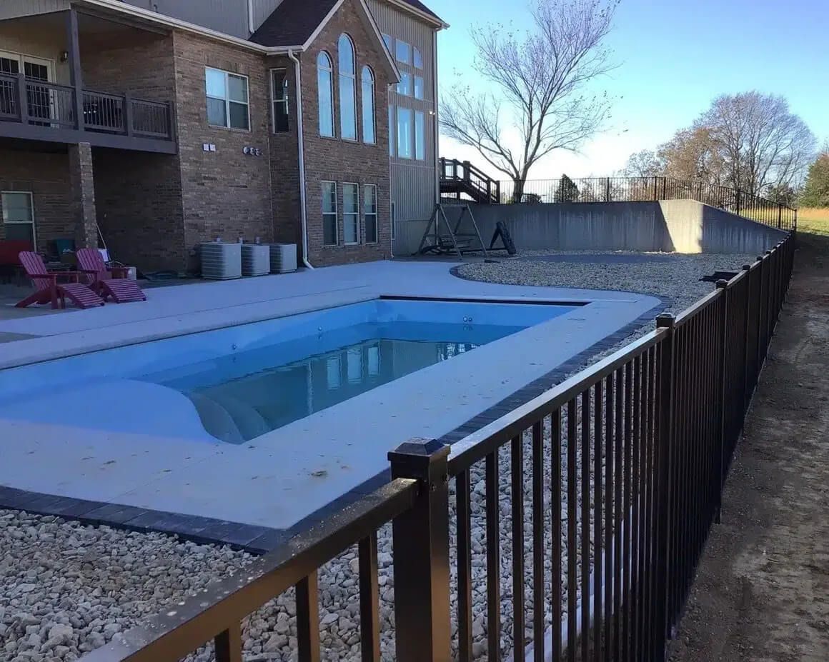 A rectangular backyard swimming pool with light gray decking, surrounded by gravel and a dark metal fence near a house.