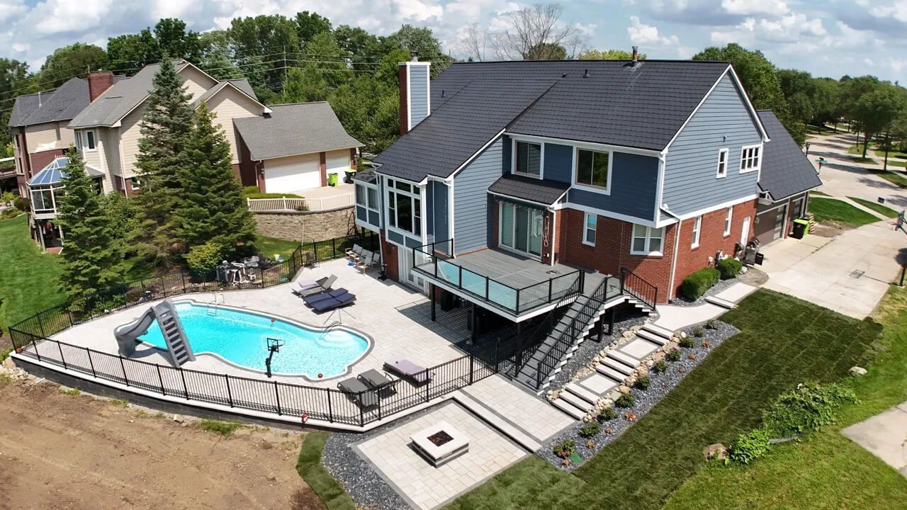 An aerial view of a blue and brick house with a backyard pool, deck, staircase, and stone fire pit area.