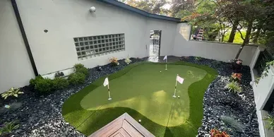 A small backyard putting green with three flagsticks, surrounded by dark decorative stone mulch and a white wall.