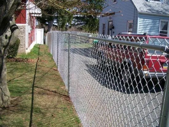 A tall chain-link fence runs along a grassy lawn next to a driveway with a red vehicle and a house.