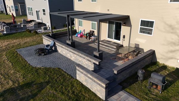 A paved stone patio with a built-in seat wall, a dark gray pergola, and outdoor dining furniture near a house.
