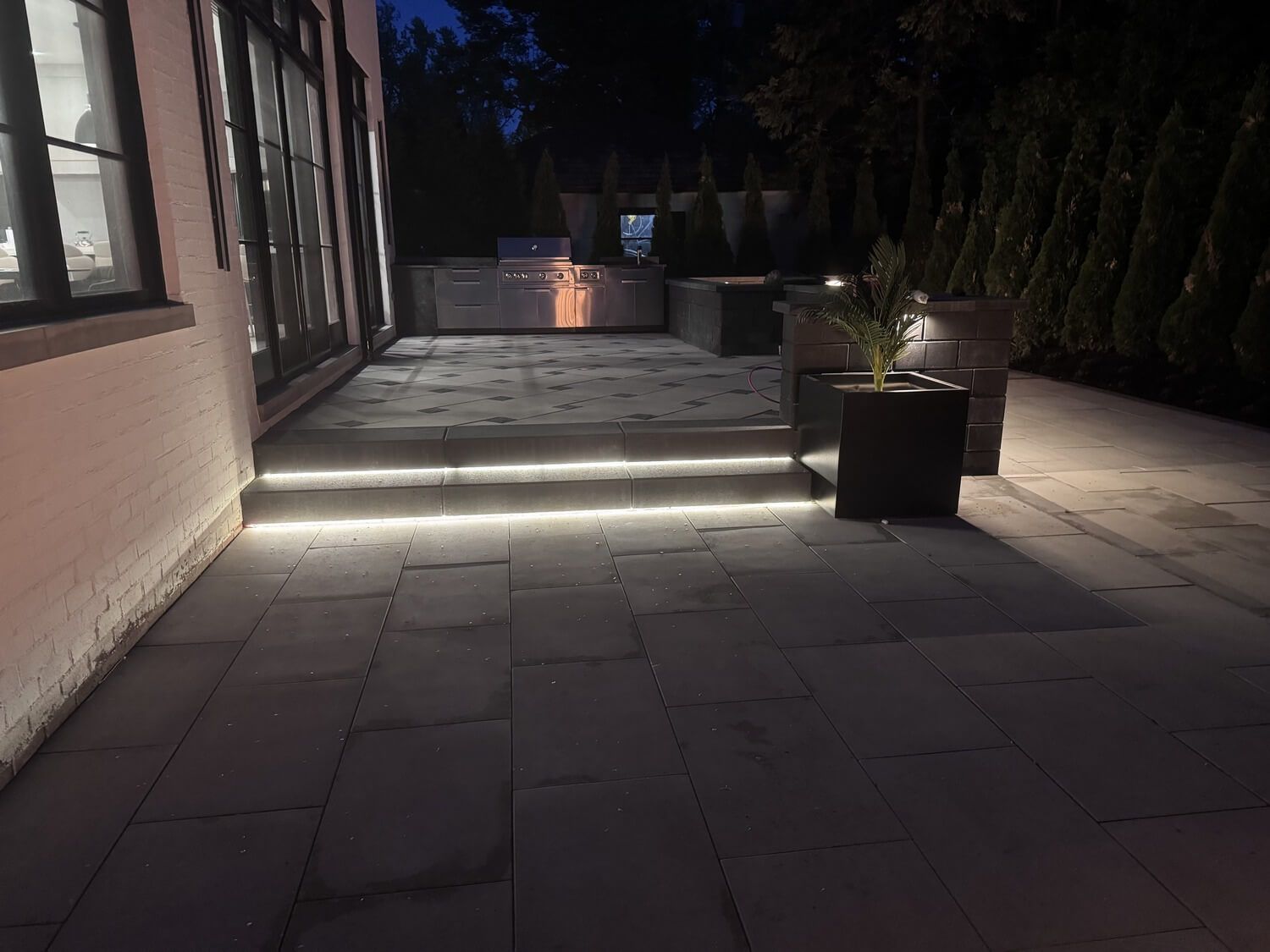A modern patio at night with stone flooring, a lit step, integrated lighting, a small planter, and an outdoor kitchen.