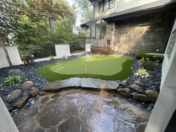A small backyard putting green with three flagsticks, surrounded by dark decorative stone mulch and a white wall.