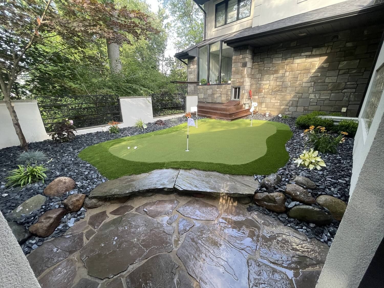 A backyard putting green with a flag sits next to a flagstone patio and rock landscaping beside a modern stone house.