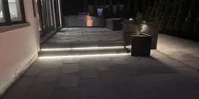 A modern outdoor patio at night with stone pavers, illuminated steps, and a lit planter beside a house exterior.