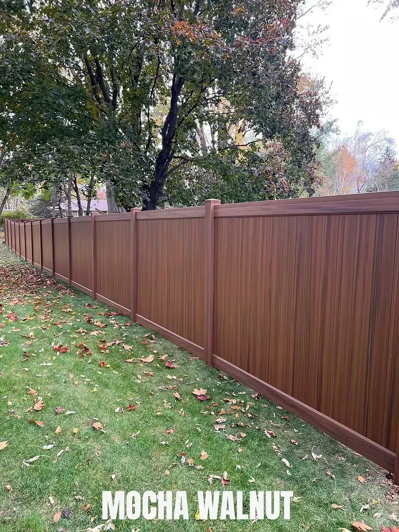 A modern privacy fence in a mocha walnut finish, installed along a grassy backyard lined with mature autumn trees.