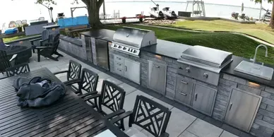 Outdoor kitchen with stainless steel grill and sink on a stone patio overlooking a lake with a table and chairs.