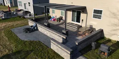 A paved stone patio with a built-in seat wall, a dark gray pergola, and outdoor dining furniture near a house.