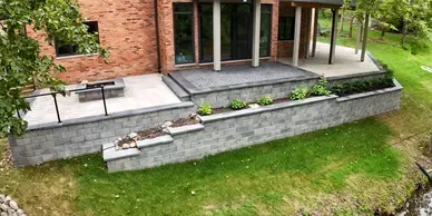 A red brick house with a multi-level stone patio, stone retaining walls, steps, and a built-in fire pit overlooking a lawn.