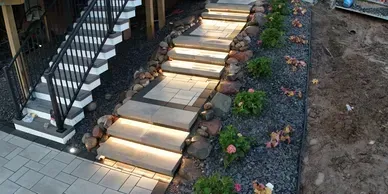 Illuminated stone steps leading outdoors next to a dark staircase and a landscape bed with gravel and small plants.