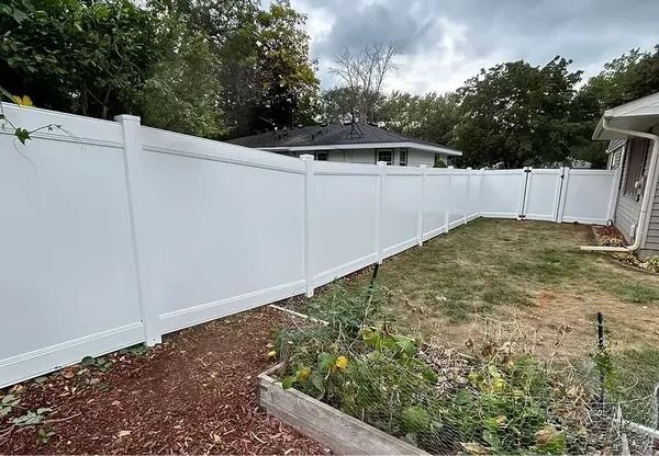 A long white vinyl privacy fence runs along a backyard, with a wooden garden bed and mulch in the foreground.