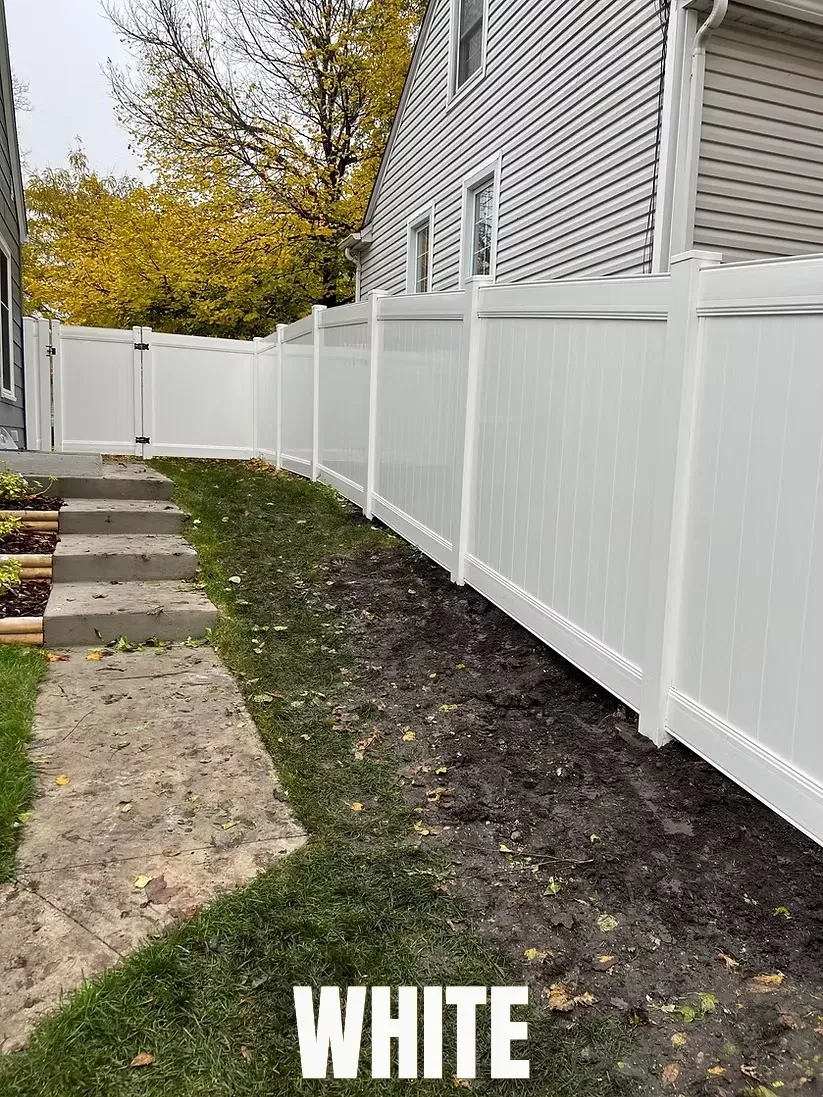 A white privacy fence stands in a yard next to concrete steps and a house, with fall foliage in the background.