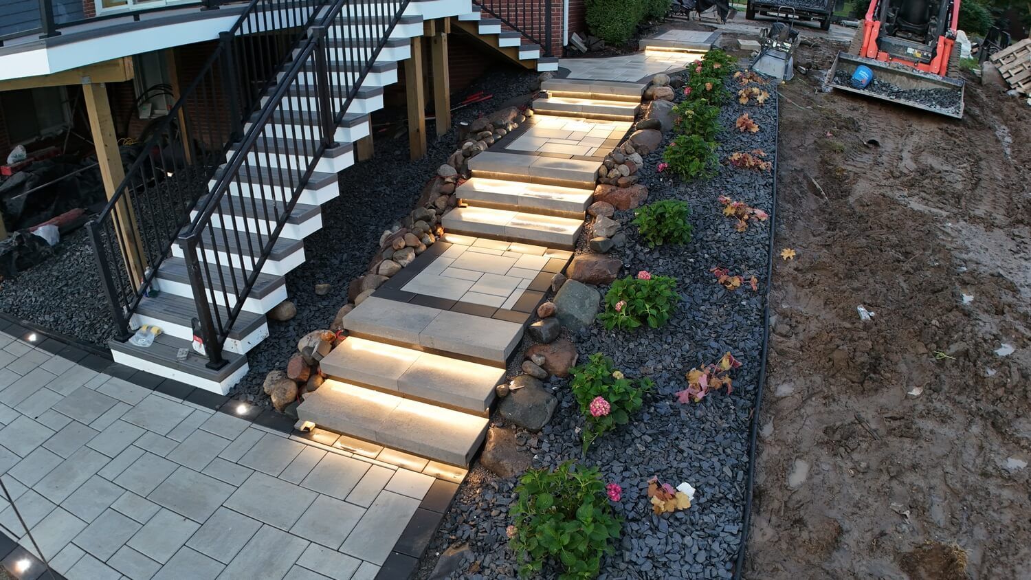 A set of light-up stone stairs leads from a patio, bordered by dark gravel and small shrubs, to a deck staircase above.