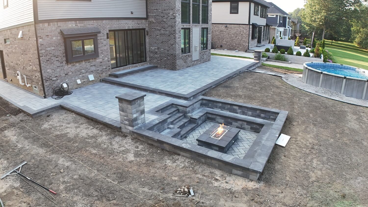 A modern brick house featuring a multilevel grey paver patio, a sunken fire pit area, and a circular pool in the backyard.