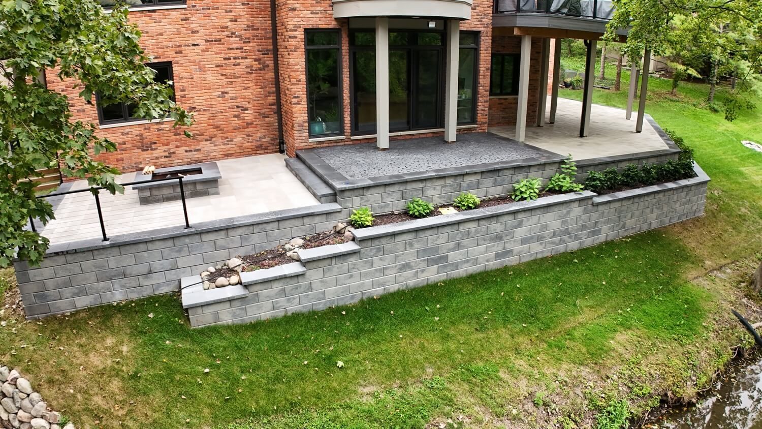 A tiered stone patio and retaining wall next to a brick house, overlooking a grassy lawn and a small creek.