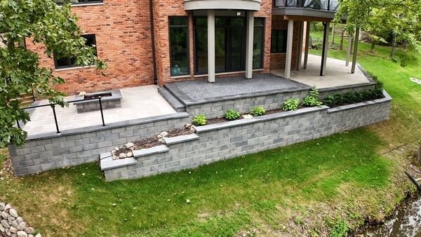 A red brick house with a multi-level stone patio, stone retaining walls, steps, and a built-in fire pit overlooking a lawn.
