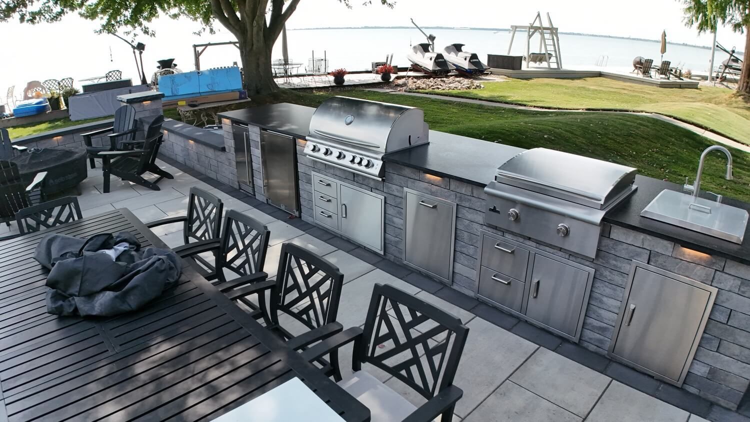 Outdoor kitchen with stainless appliances, stone counters, and a dining set on a stone patio overlooking a lake.