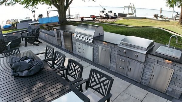 Outdoor kitchen with stainless steel grill and sink on a stone patio overlooking a lake with a table and chairs.