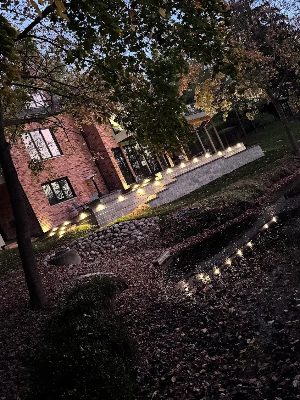 A brick house at dusk, illuminated by a string of warm fairy lights along a gravel path and through a leaf-covered yard.