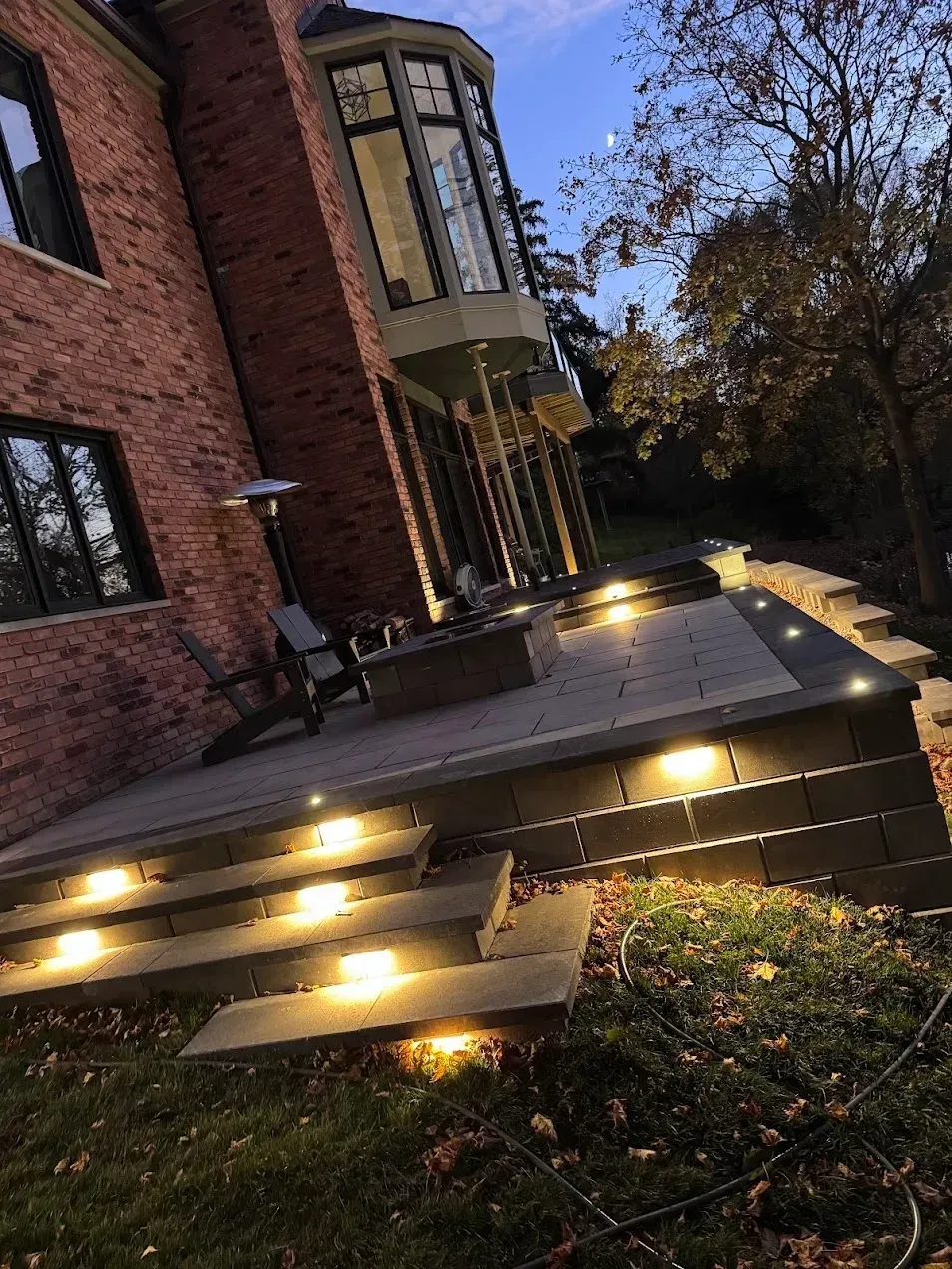 A brick house with a raised stone patio illuminated by warm step and perimeter lights at twilight.